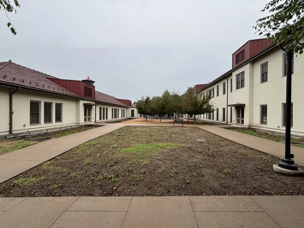 A before photo of the large ground space in the center of the residence hall unattended with patched grass and plants.